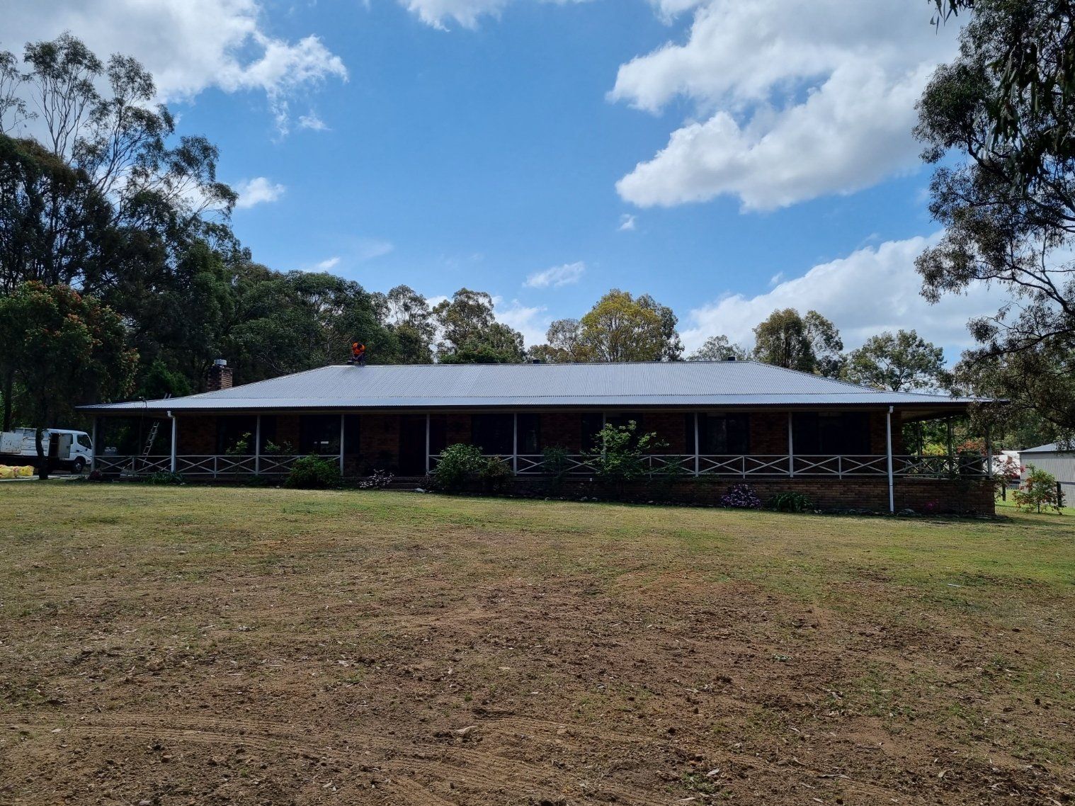 A single-story ranch house with a wrap-around veranda and dark roof, set on a grassy lot against a blue, cloudy sky.