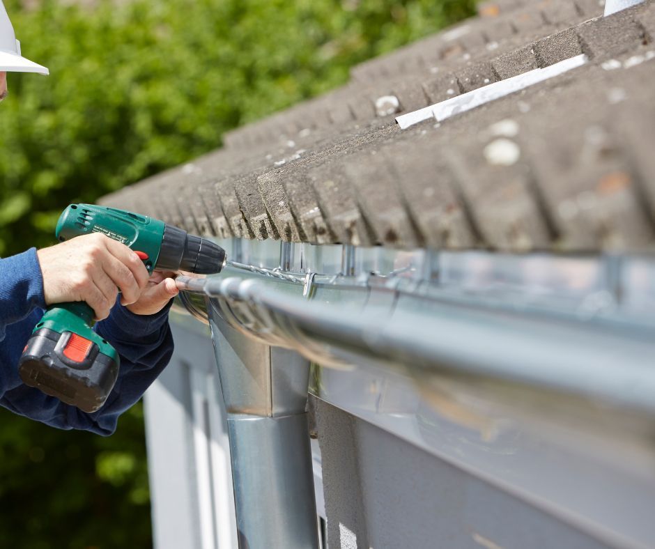 A person wearing a white hard hat uses a green electric drill to secure a metal gutter to the side of a tiled roof.