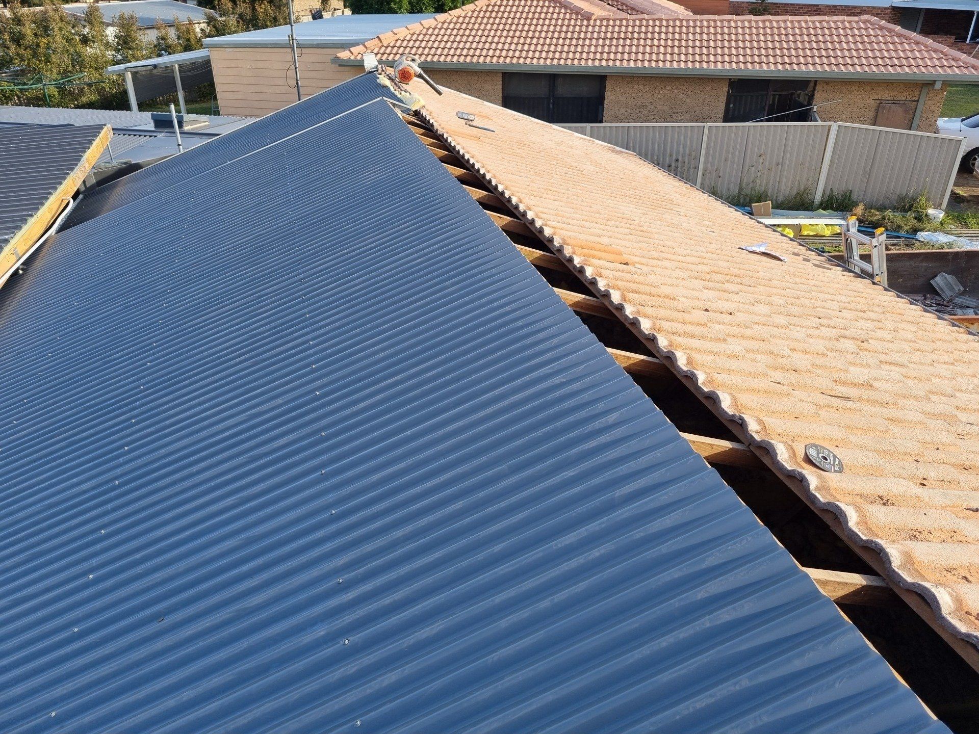 A partially reroofed house with a new dark corrugated metal roof section next to an older tan tiled section.