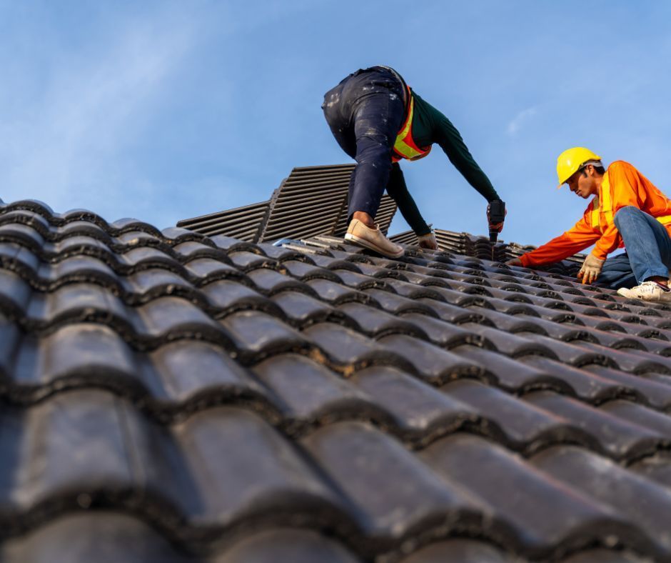 Two workers in high-visibility clothing and hard hats install tiles on a residential roof under a clear blue sky.