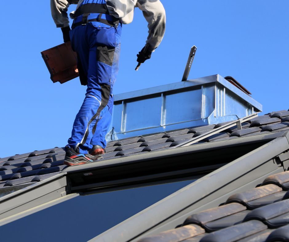 A worker in blue workwear walks on a tiled roof near a metal chimney and skylight against a clear blue sky.