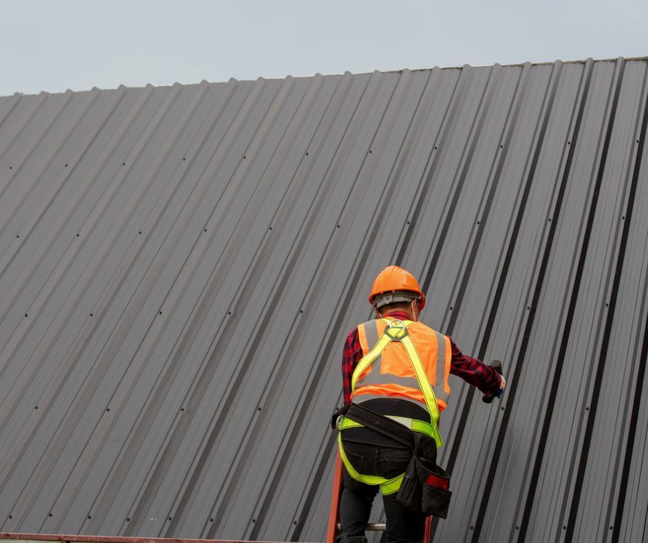 A worker in an orange hard hat and safety vest stands on a ladder working on a dark gray corrugated metal roof.