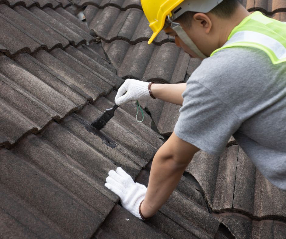 Worker in hard hat inspecting roof tiles with a tool, wearing gloves and safety vest.
