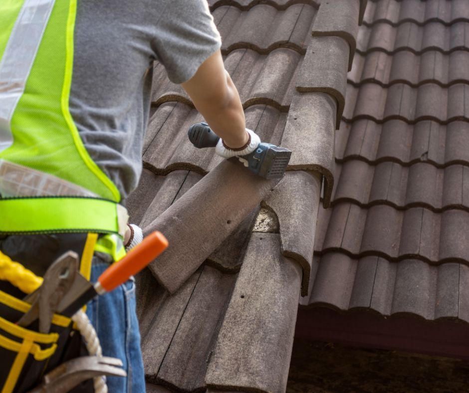 A worker in a high-visibility vest and tool belt repairs brown roof tiles on a sloped roof.