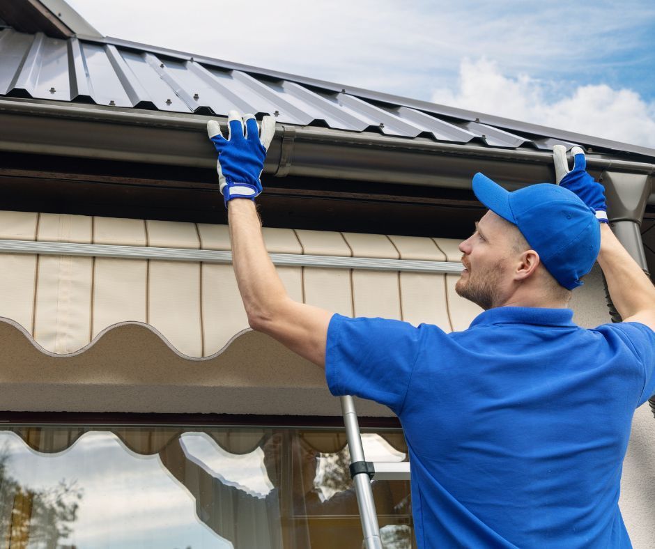 Worker in blue uniform inspecting and cleaning a house roof gutter with a ladder nearby
