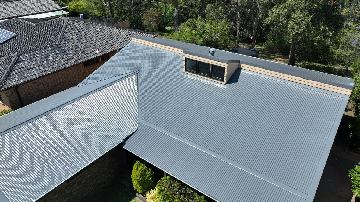 An aerial view of a dark grey metal roof with a skylight and a raised ridge section, surrounded by trees and a house.