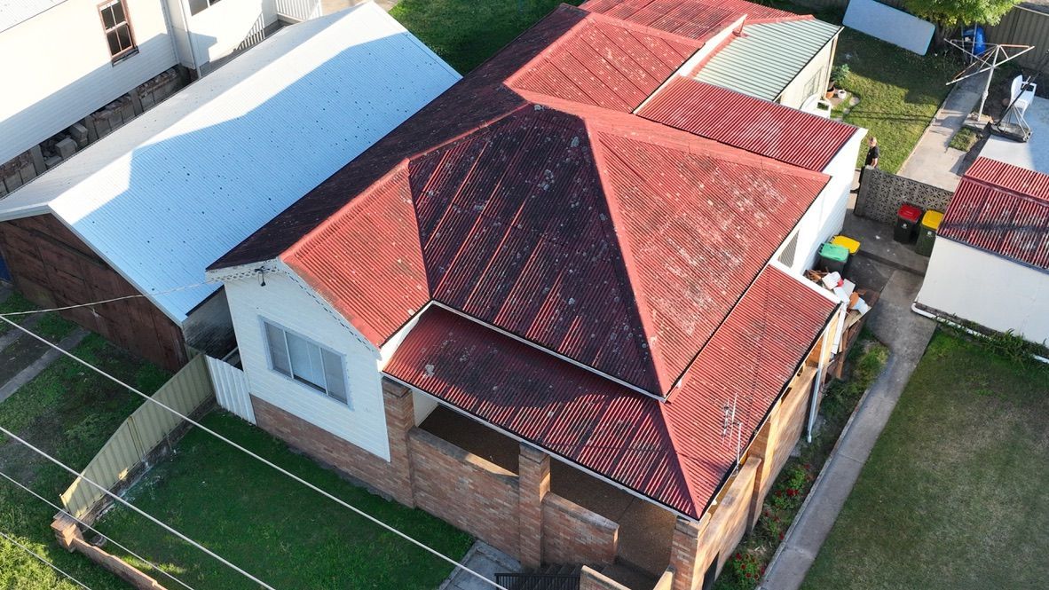 Aerial view of a residential brick house featuring a dark red, multi-pitched metal roof and a small front porch.