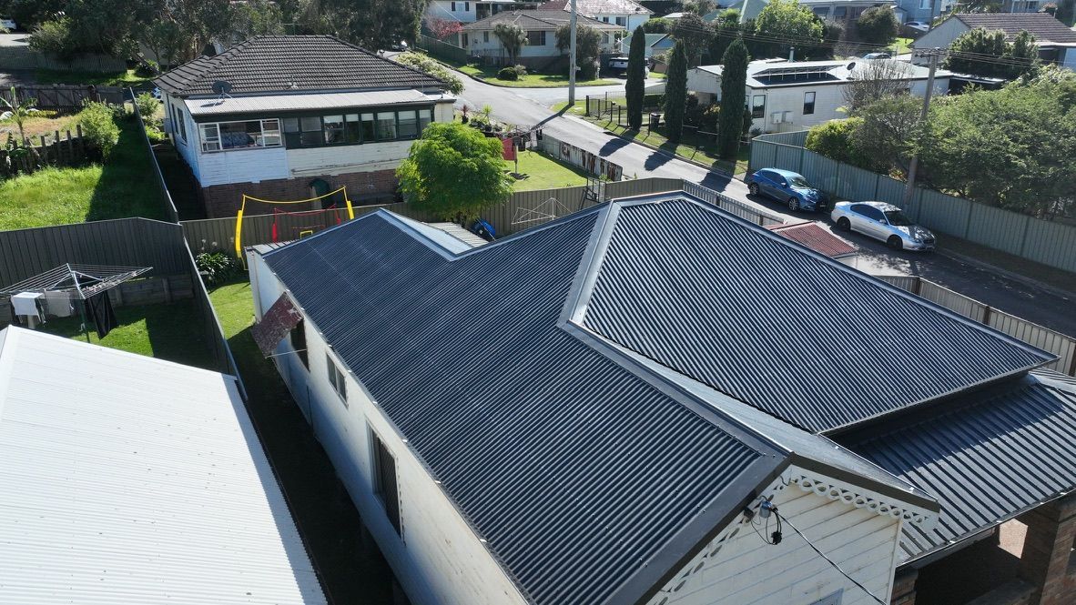 An aerial view of a residential street featuring a house with a dark, ridged metal roof, surrounding yards, and vehicles.