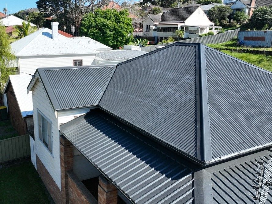 An aerial view of a dark grey corrugated metal roof on a suburban house with a brick base and adjacent white homes.