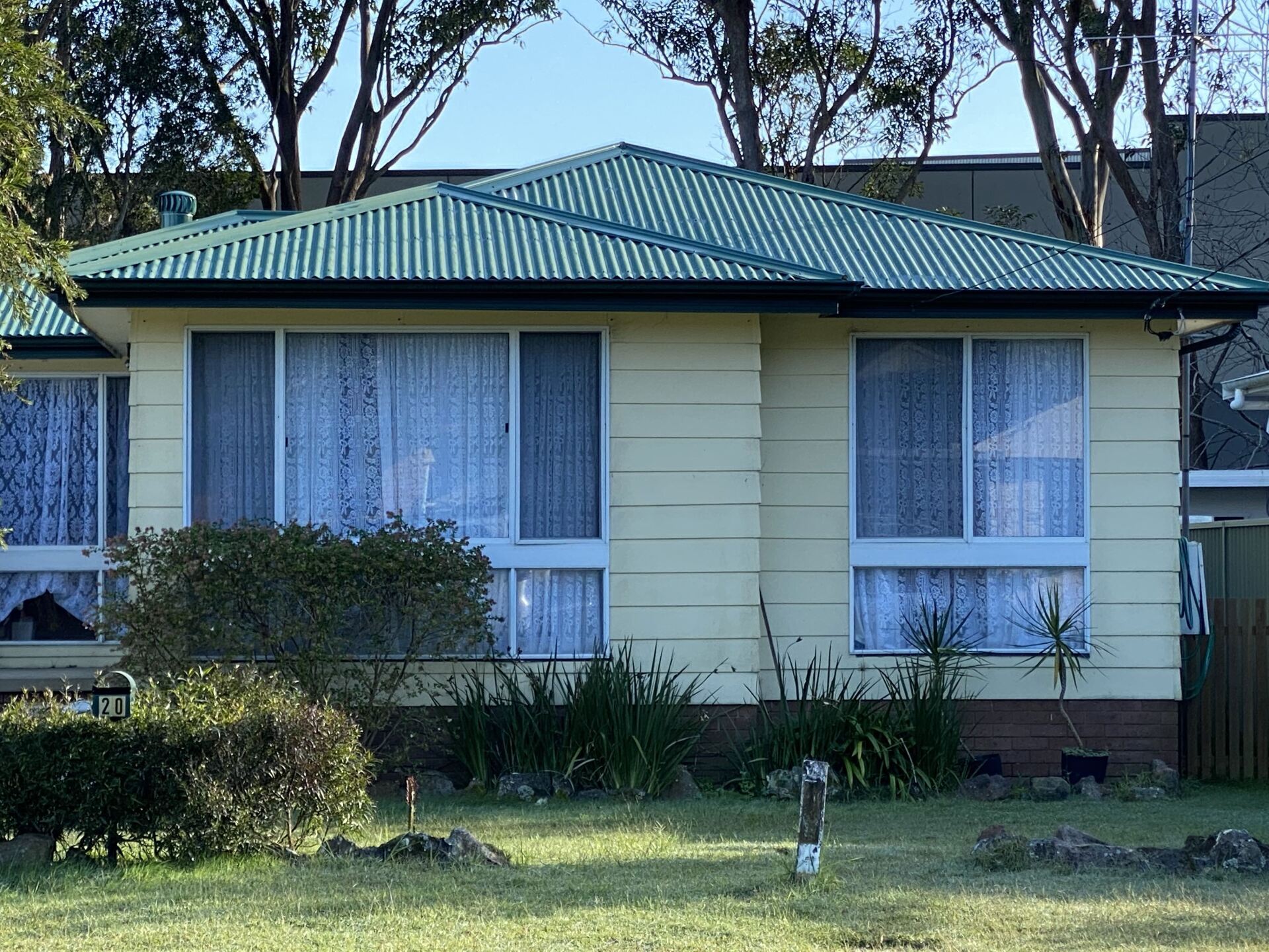 A single-story, yellow suburban house with a green corrugated metal roof, surrounded by lawn and trees under a blue sky.