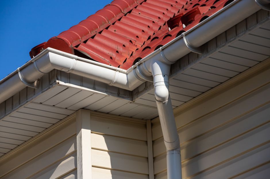A close-up of a house exterior featuring a red tiled roof, white gutters, a downspout, and light beige siding.