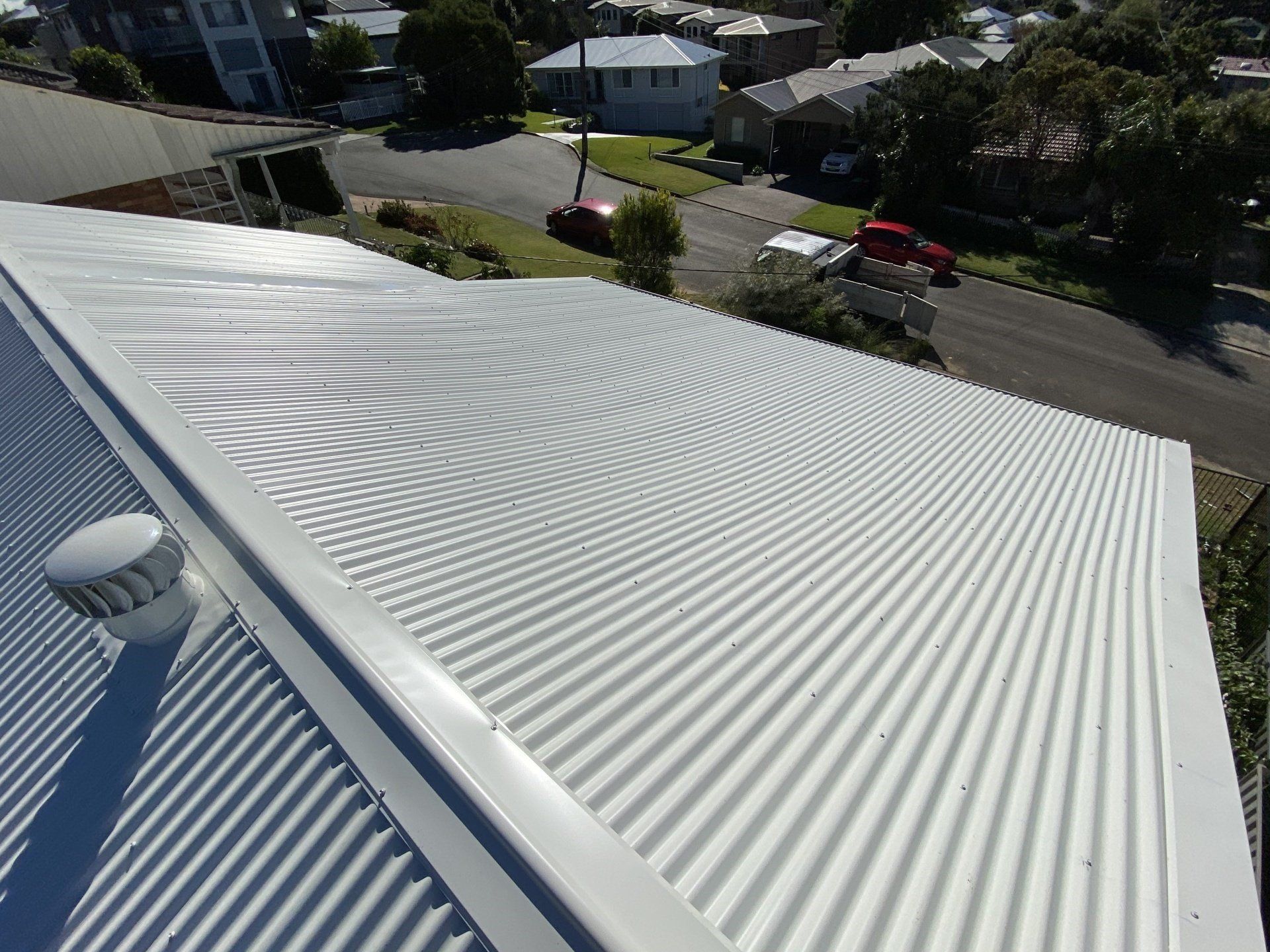 A high-angle view of a white, corrugated metal roof with a vent, overlooking a suburban street with houses and cars.