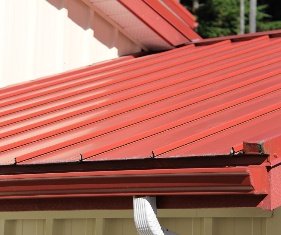 A red standing seam metal roof with a gutter and downspout on a building with beige walls.