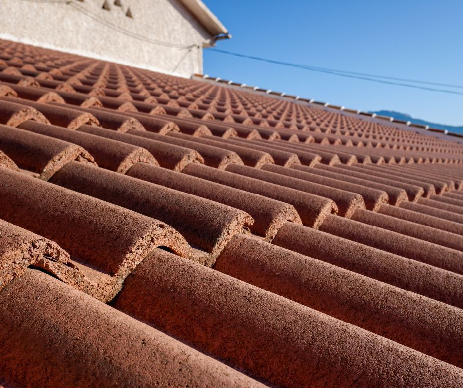 A close-up view of a sloping roof covered in textured, reddish-brown terracotta tiles under a clear blue sky.