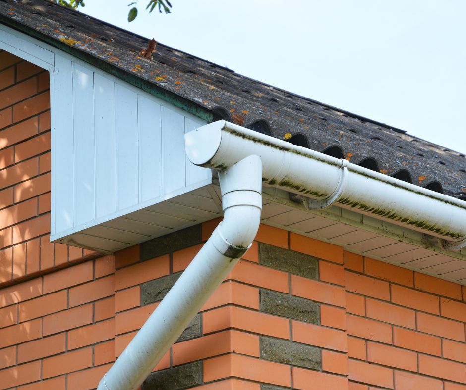 White plastic gutter and downspout attached to the corner of a brick building with a dark tiled roof and soffit.