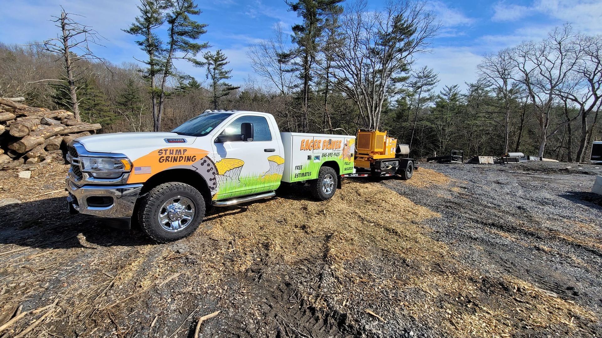 White utility truck with orange and green graphics parked on a gravel lot beside a trailer, near trees and brush.