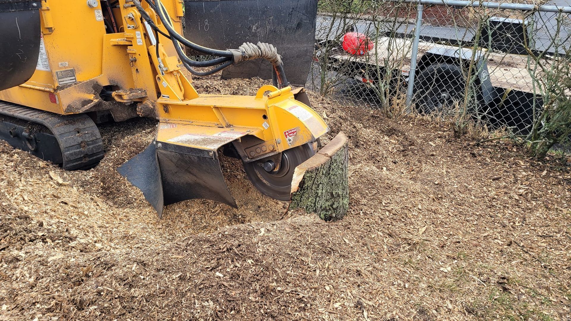 Yellow backhoe clearing brush and debris beside a fence in a wooded area.