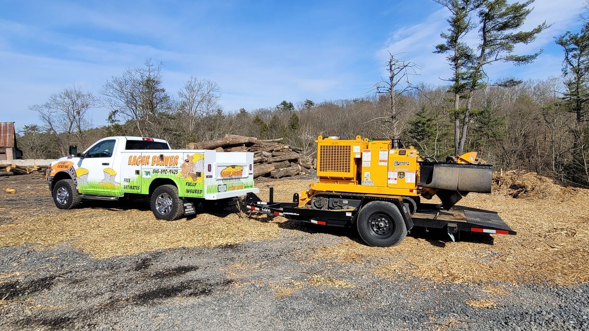 Two utility trucks with equipment trailers parked on gravel in a wooded area under clear skies