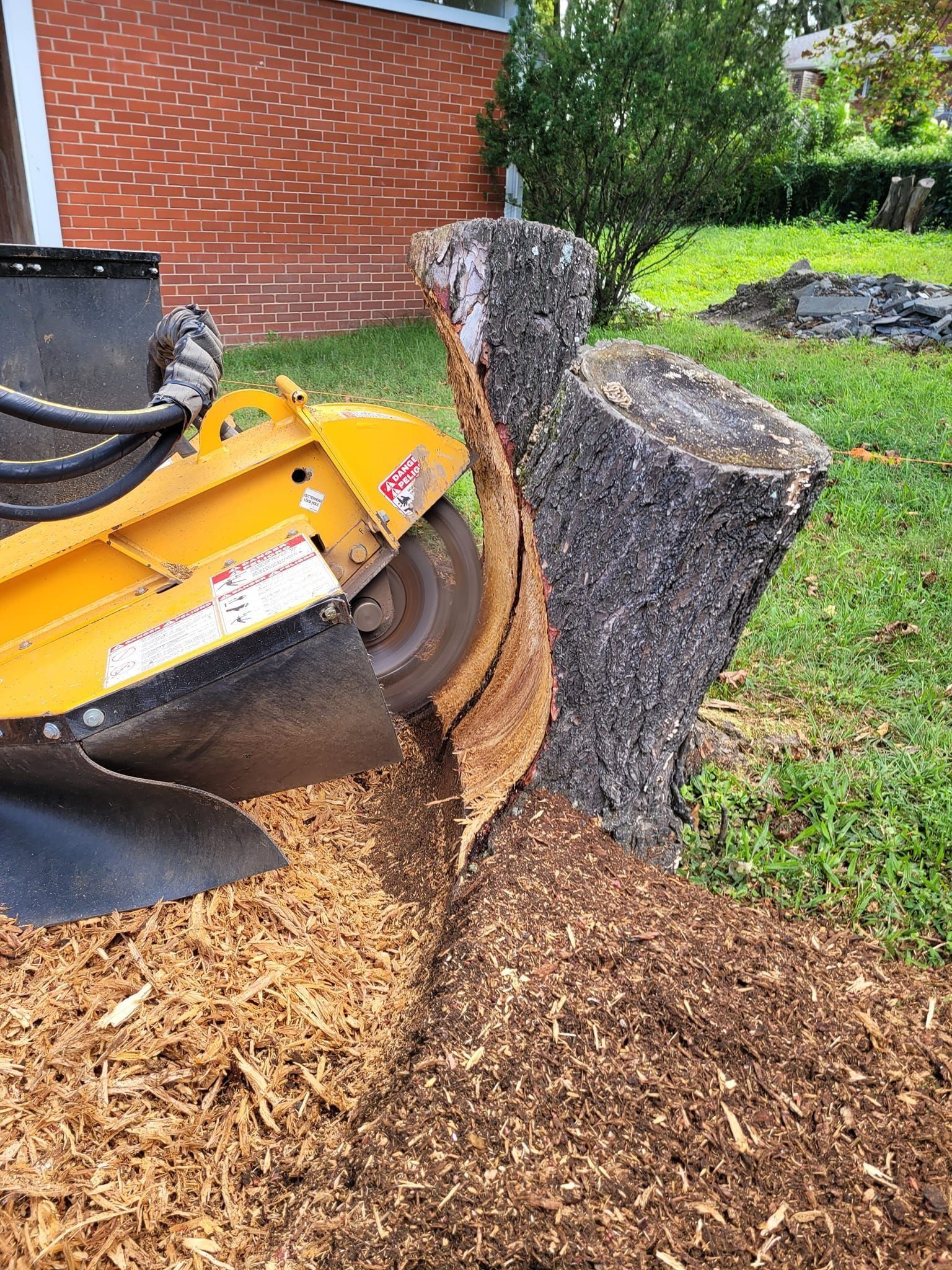 Yellow mulching machine grinding a tree stump beside a brick house, with wood chips on the ground
