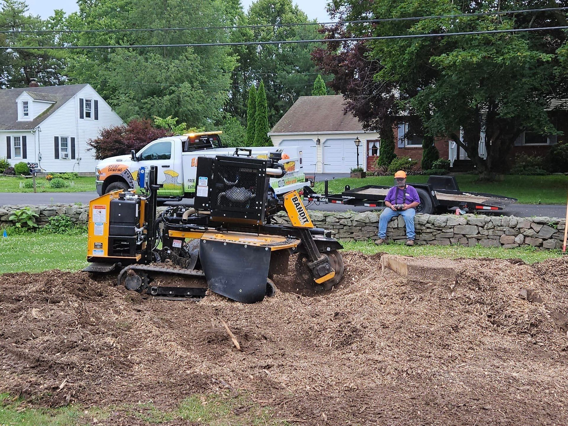 Yellow skid-steer working on dirt beside a stone wall, with a person sitting nearby in a yard.