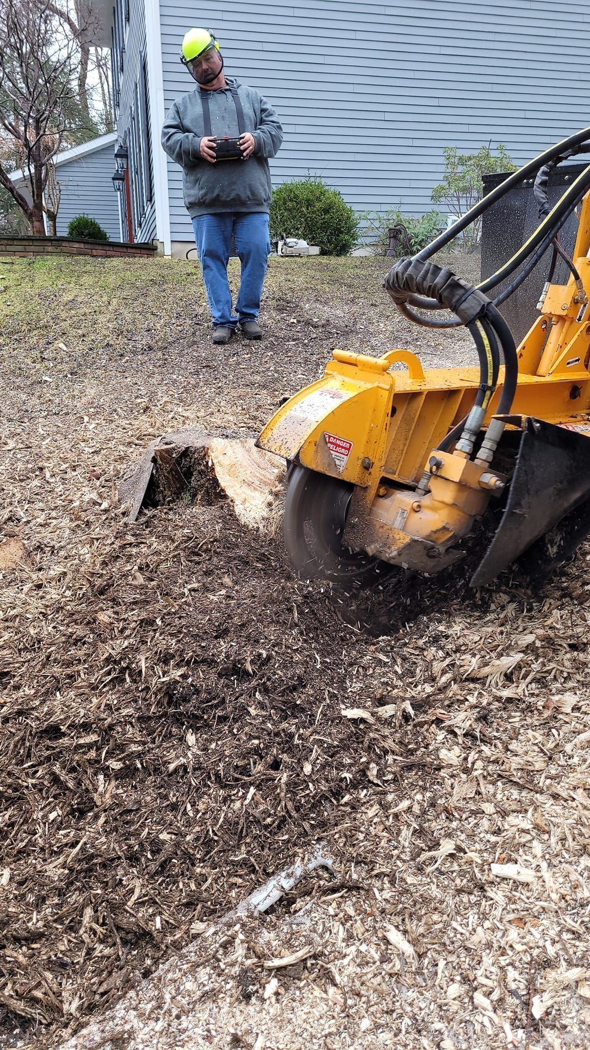 Yellow wood chipper mulching a pile of bark mulch while a worker stands nearby outside a building.
