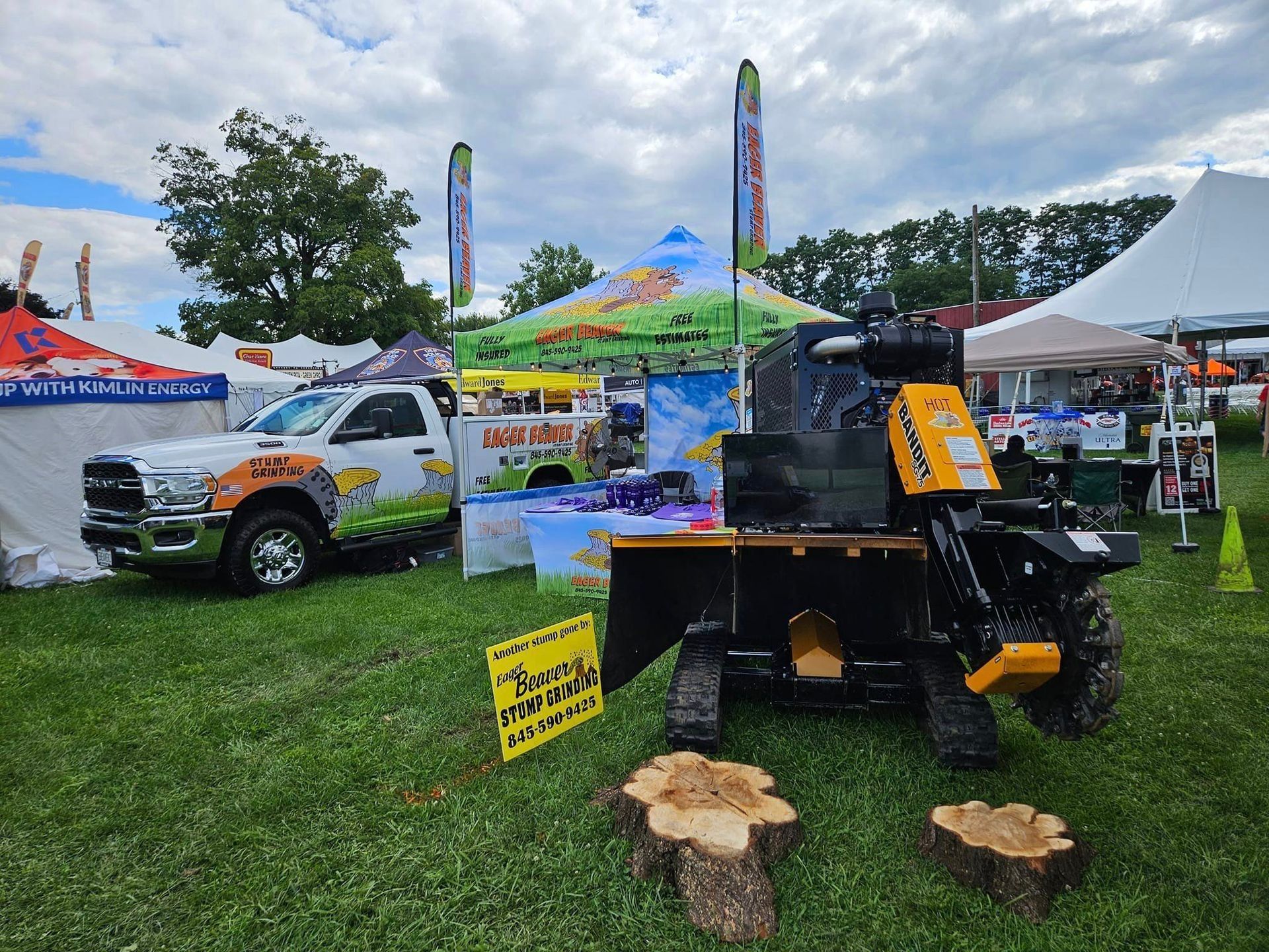 Outdoor BBQ setup with a black smoker grill, logs, and colorful vendor tents at a festival