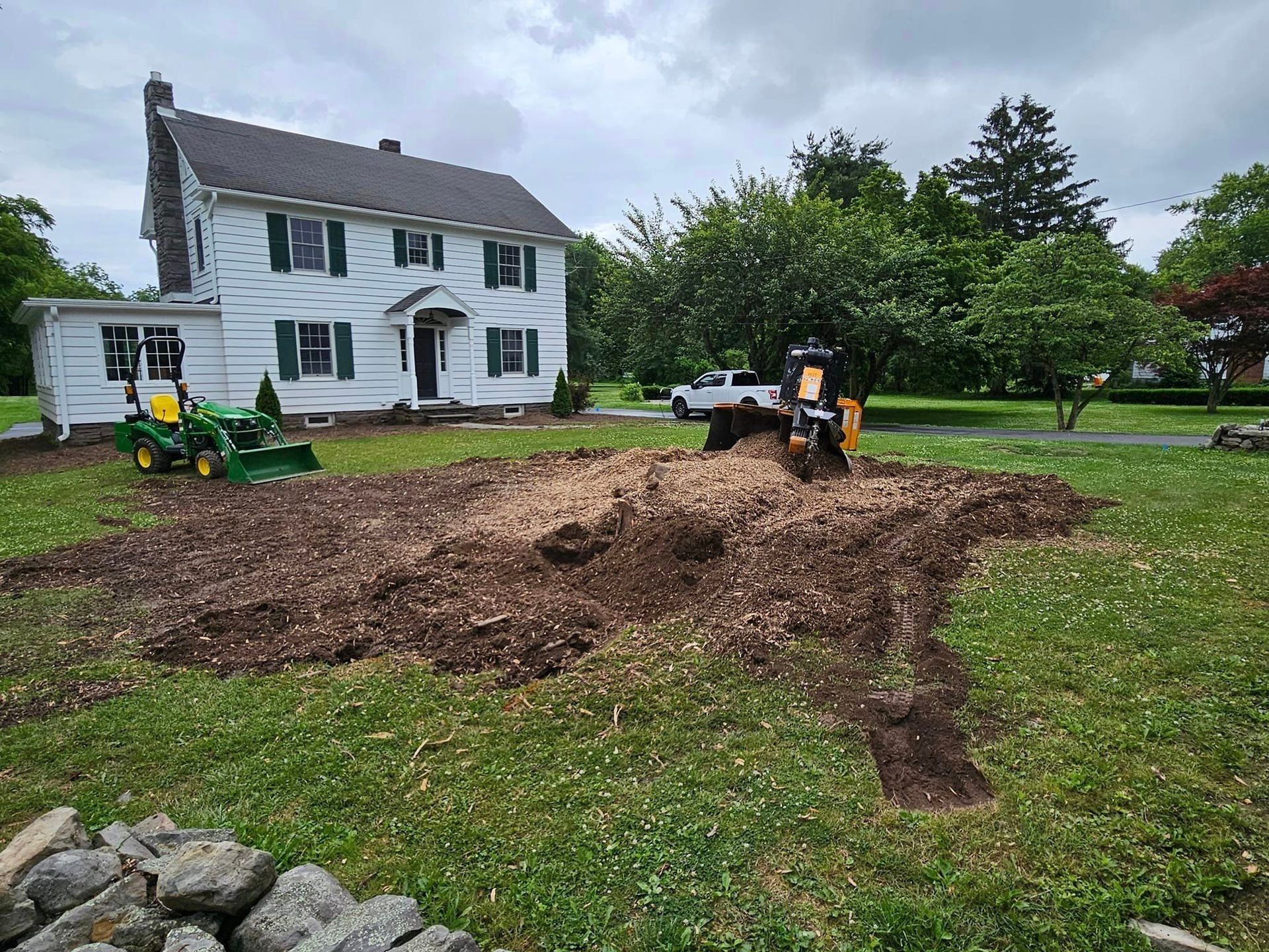 Backyard excavation with a small excavator and dirt pile beside a white house and lawn
