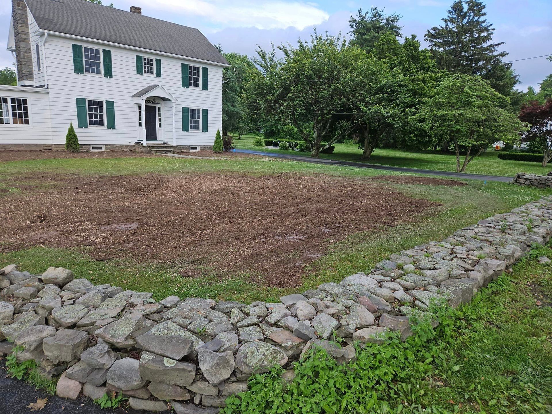 Stone-bordered garden bed in front of a white house with green shutters and a lawn.
