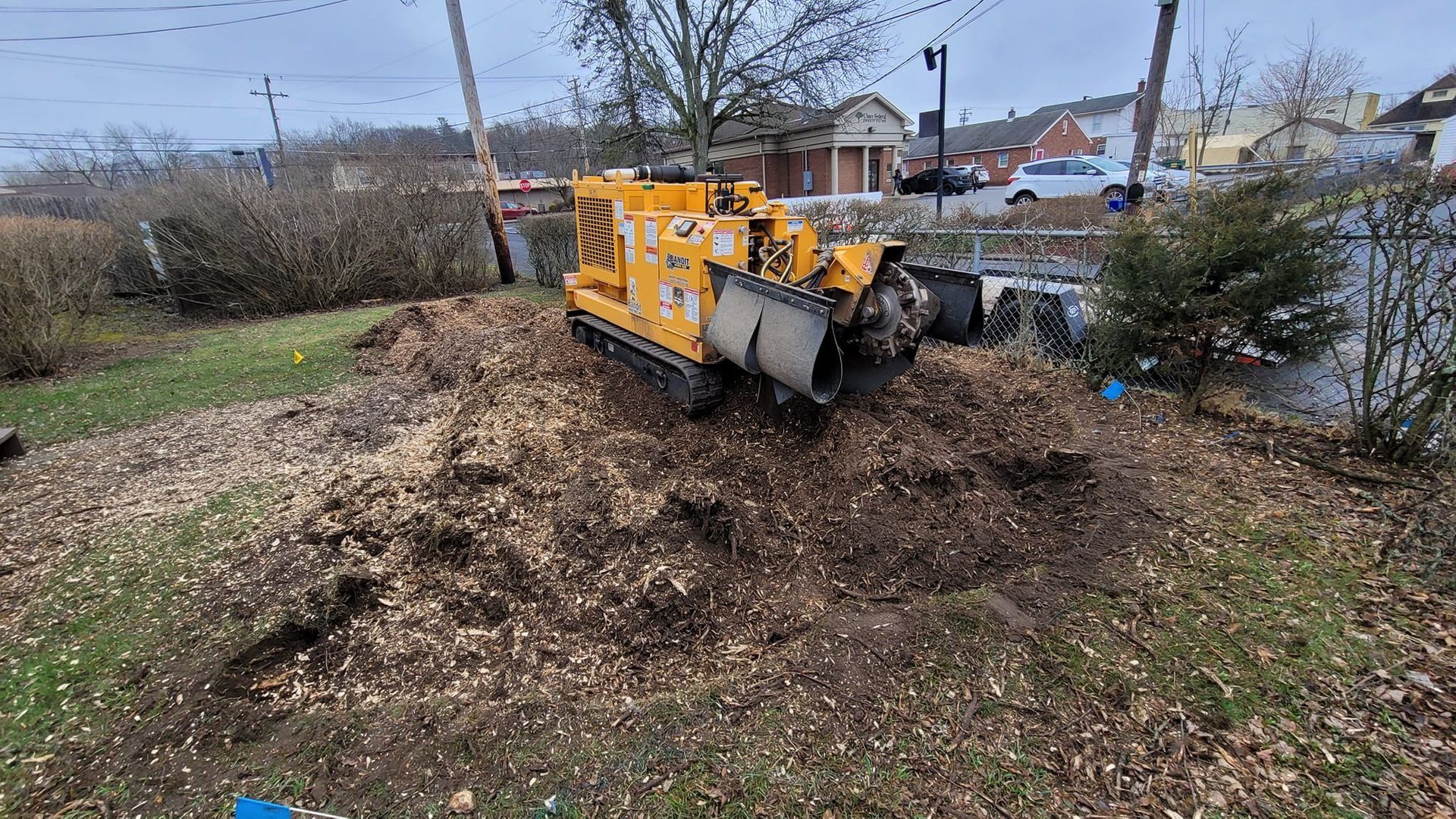 Yellow skip loader piled with brush on a grassy roadside near a stream, under cloudy skies