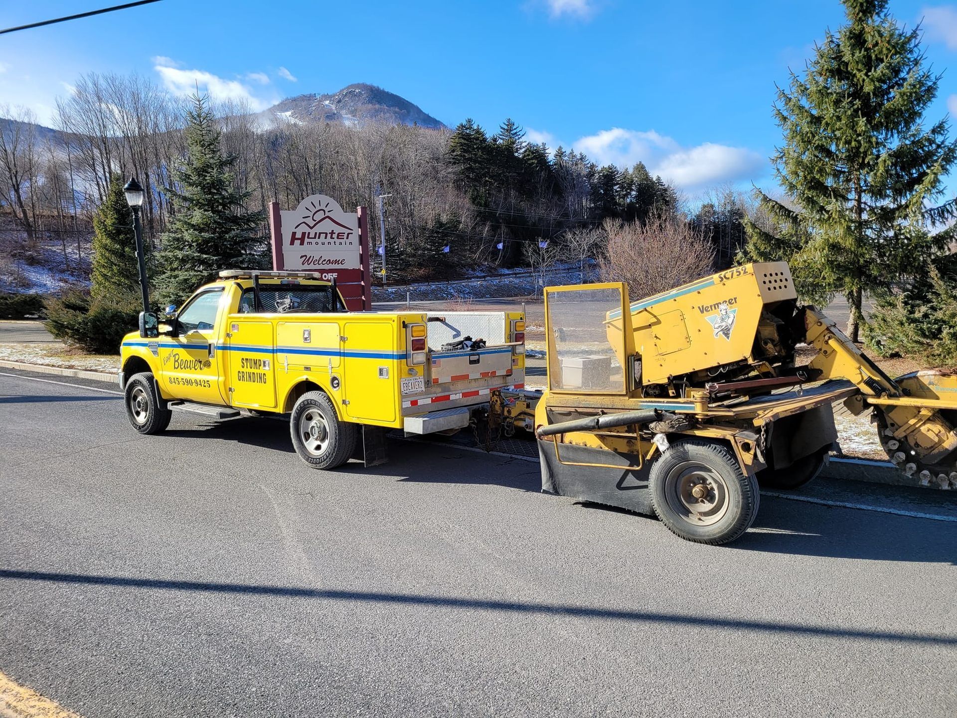 Yellow utility truck towing a yellow trailer with equipment on a mountain roadside under a blue sky