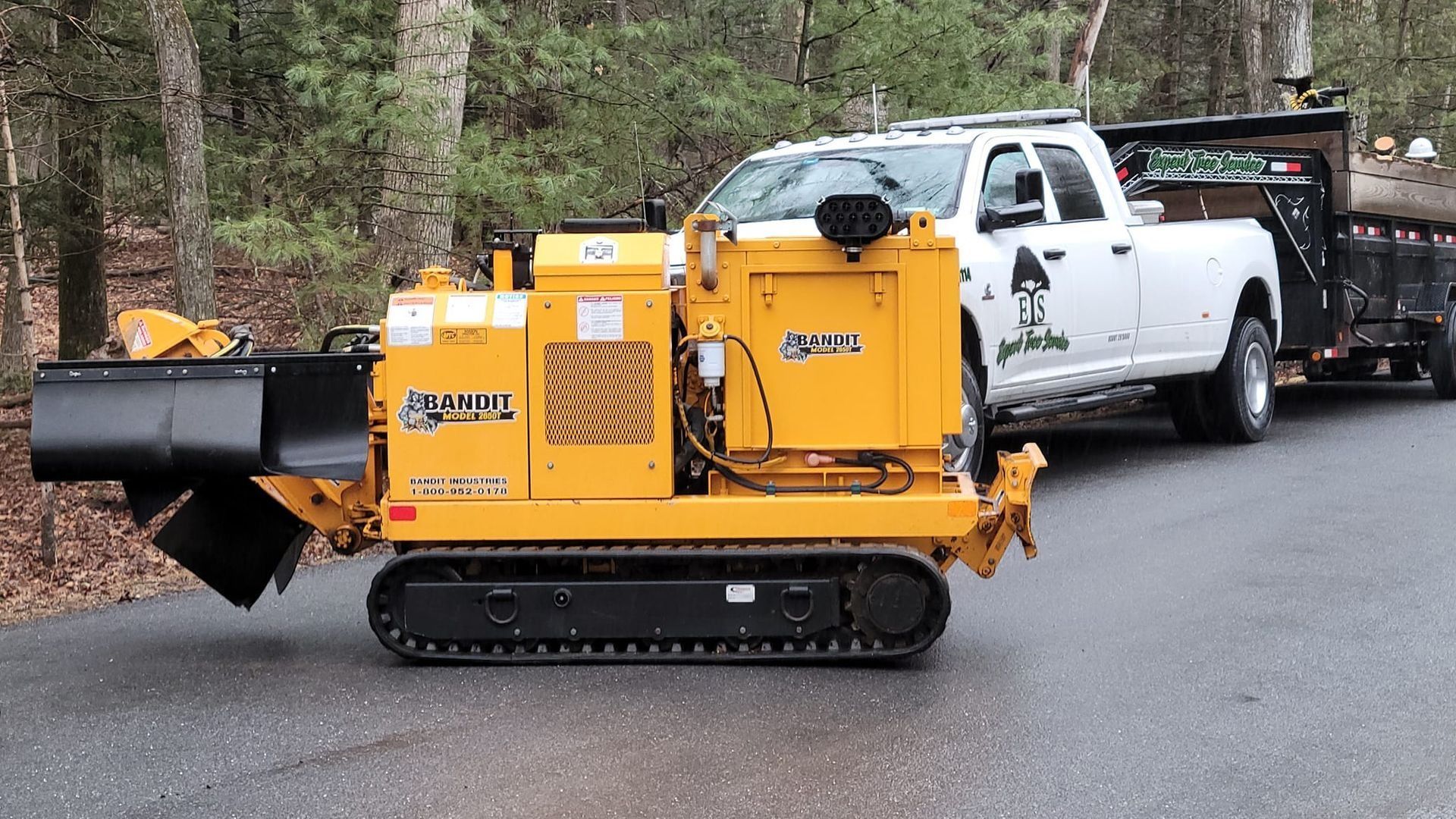 Yellow tracked stump grinder parked on a road beside a white pickup truck in a wooded area
