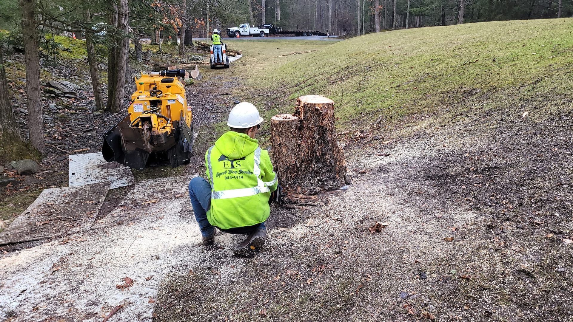 Worker in a neon jacket kneels near a stump beside a yellow ATV on a muddy trail in a wooded area