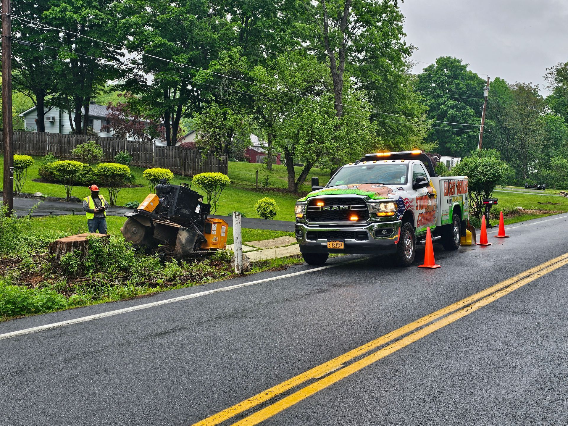 Roadside work zone with a utility truck, traffic cones, and a worker clearing debris beside a wet road.