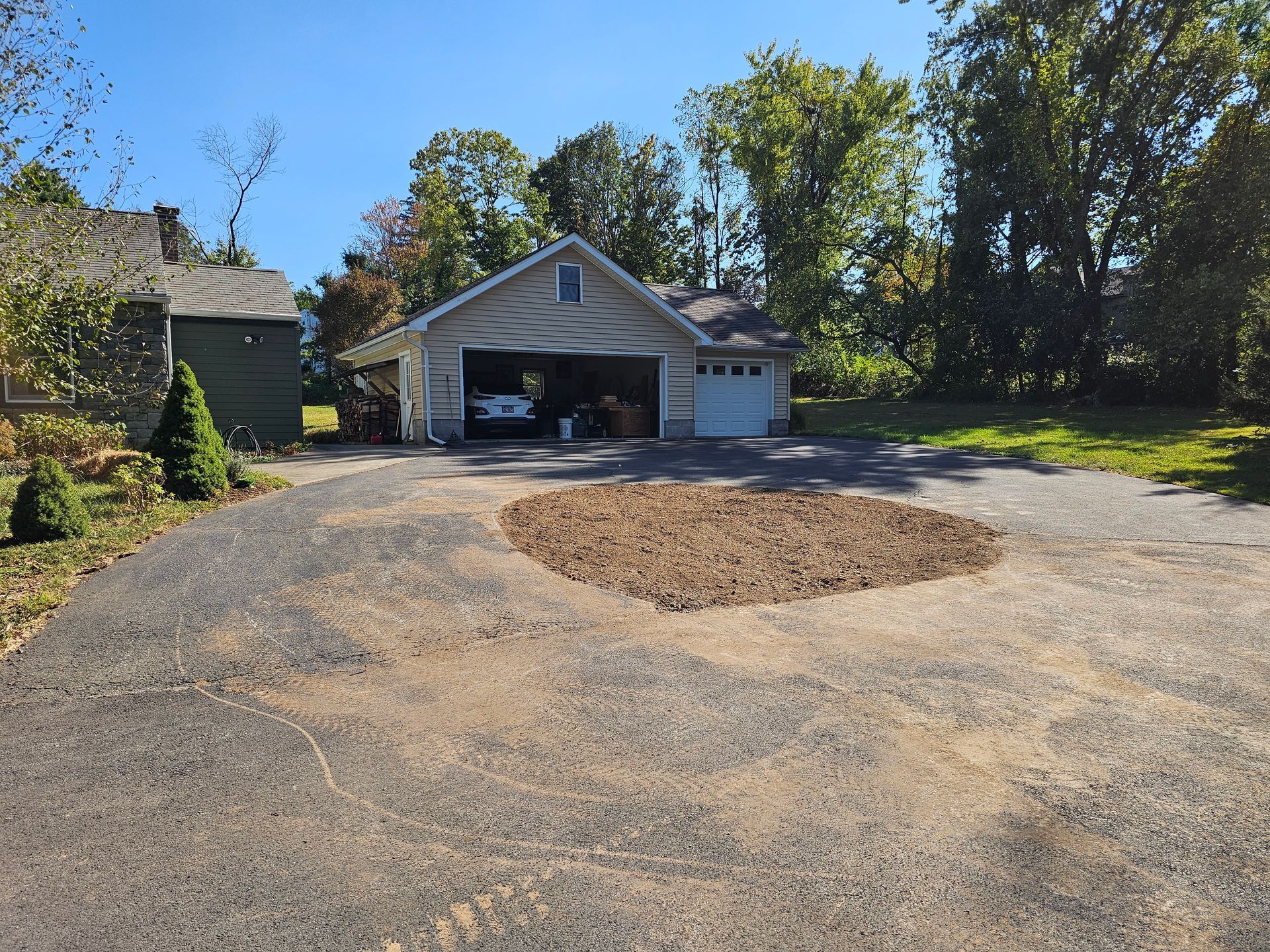 Gravel driveway leading to a garage with a circular mulch bed in front, surrounded by trees and autumn foliage