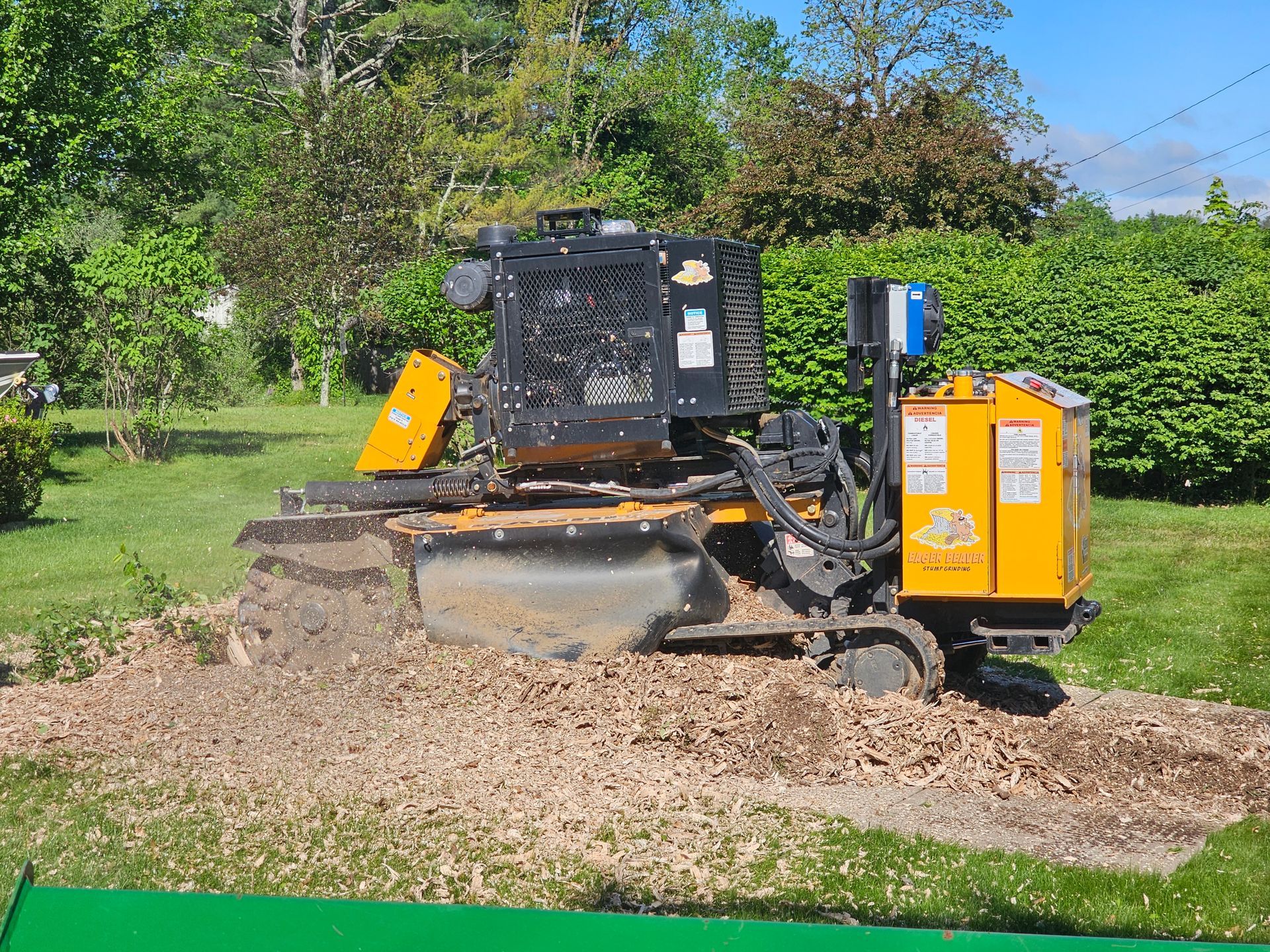 Yellow and black construction machine parked on gravel in a grassy park with trees and hedges