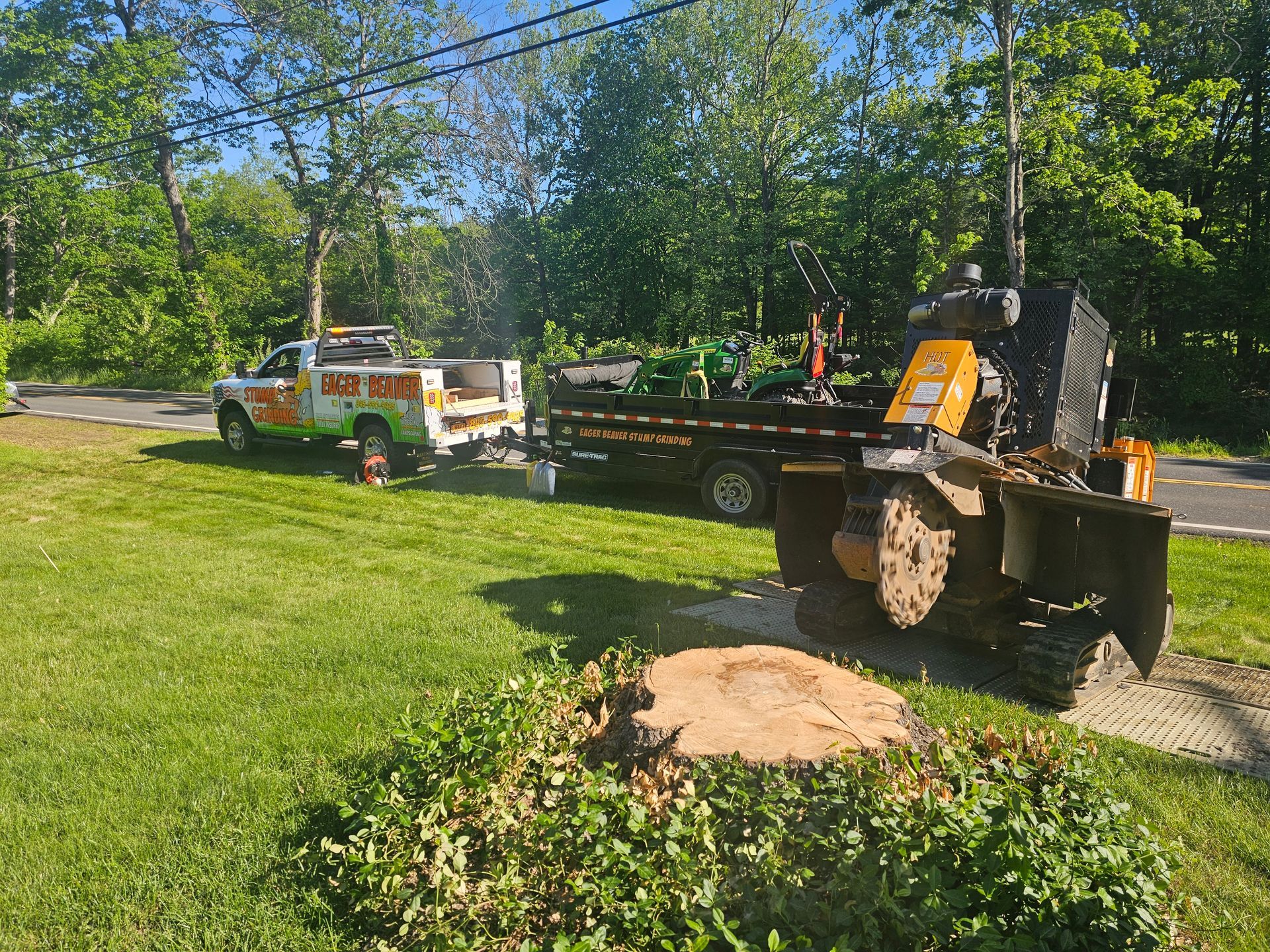 Truck and flatbed trailer with excavator on a grassy roadside under trees