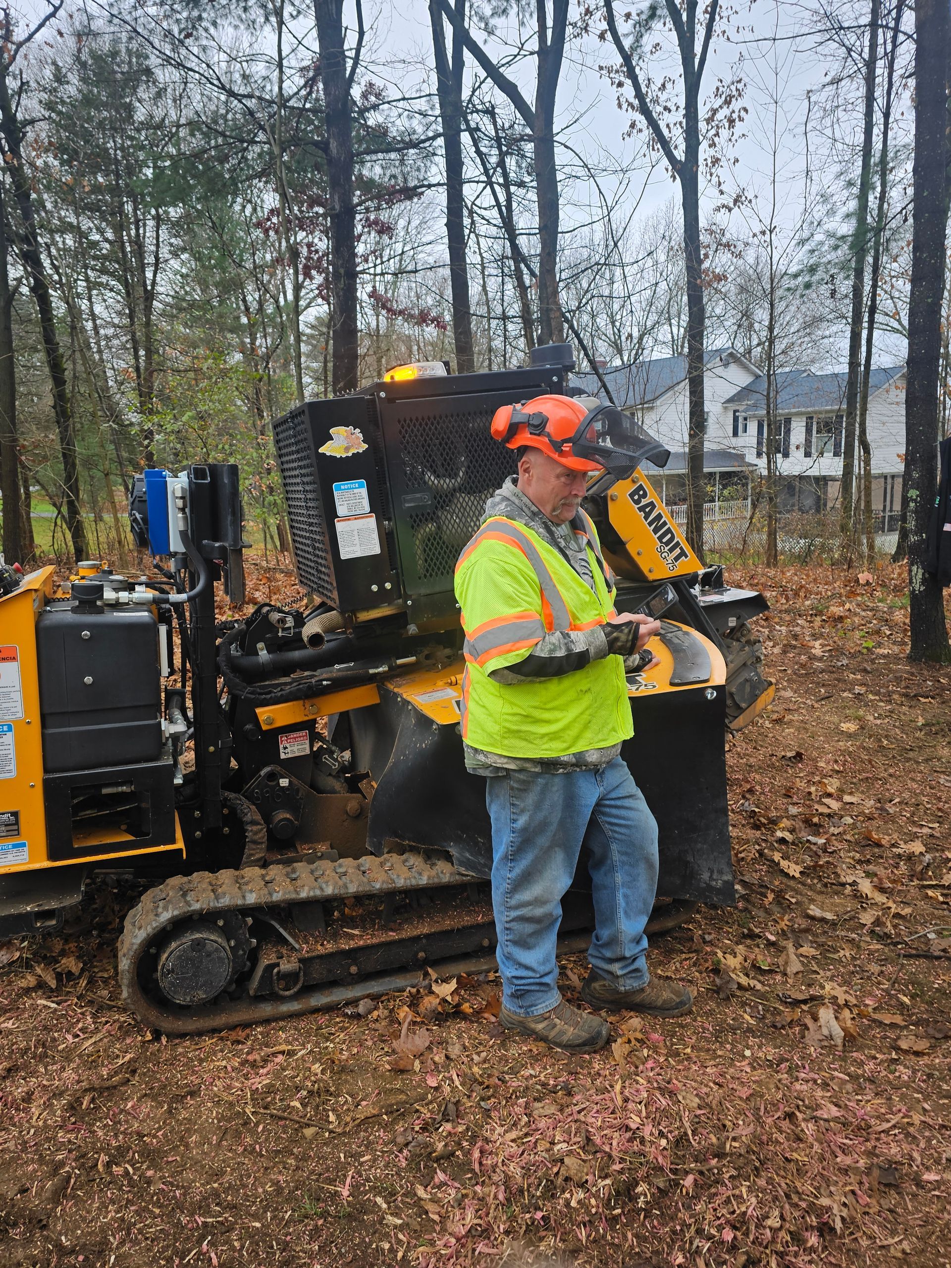 Worker in orange hard hat stands beside a yellow tracked machine in a wooded area.