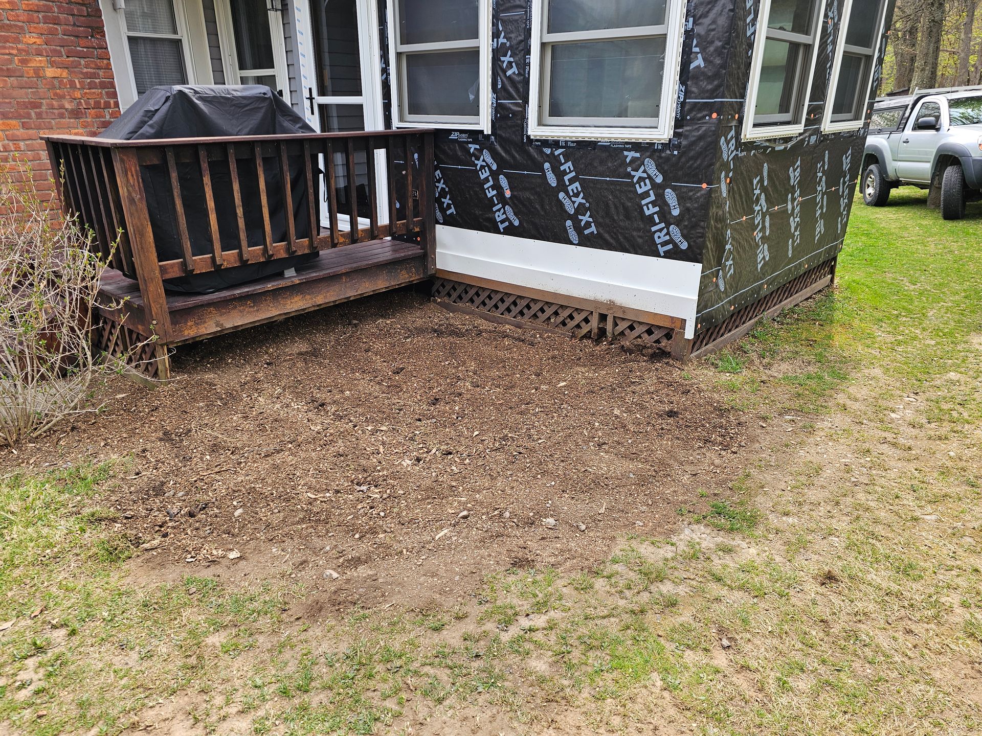 Small wooden porch on a dark house, bordered by fresh mulch and a grassy yard with a car nearby.
