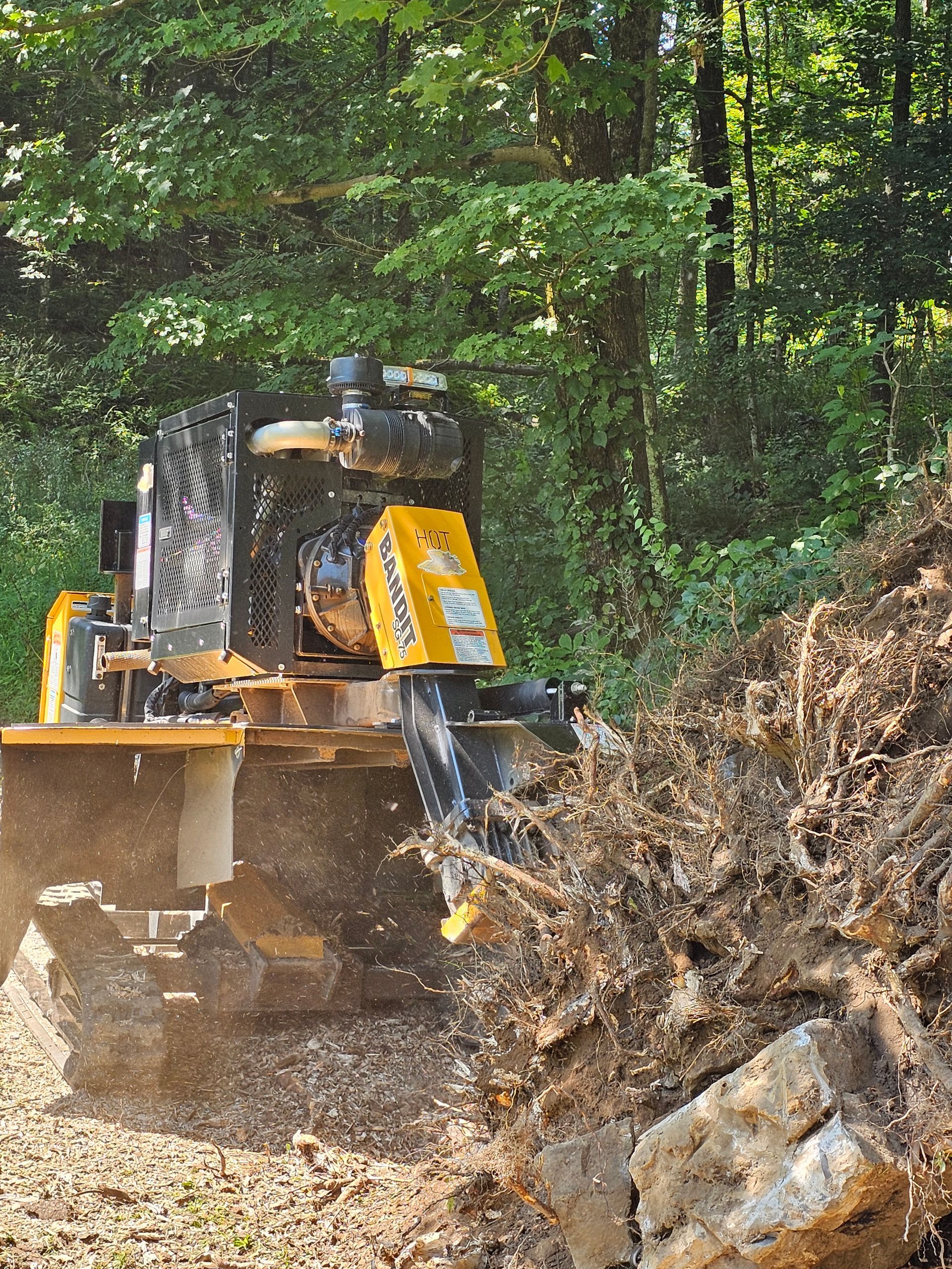 Yellow bulldozer clearing dirt on a wooded hillside, pushing debris beside a rocky embankment.