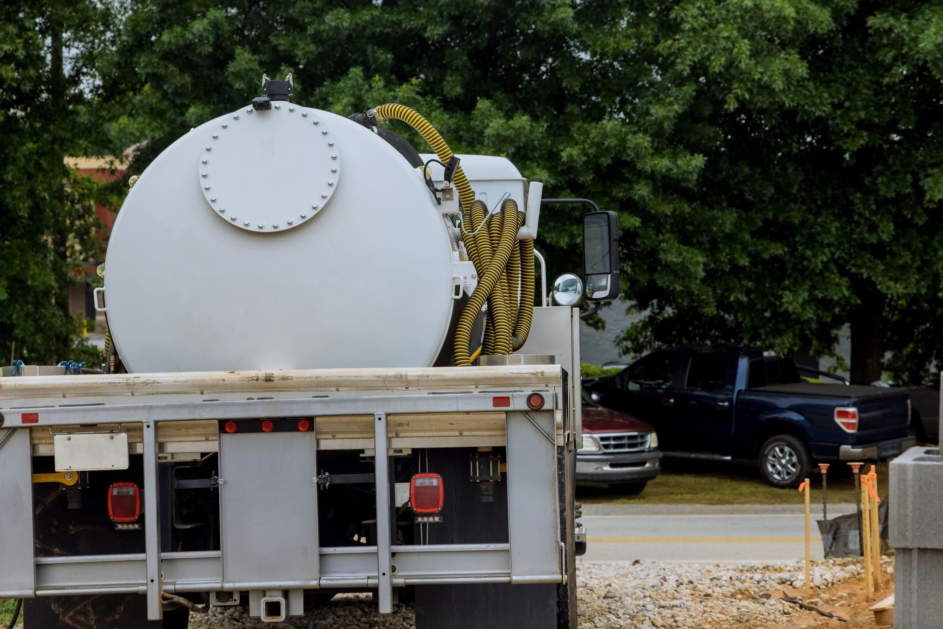 White industrial tank on a septic truck parked by a road with trees and cars in the background