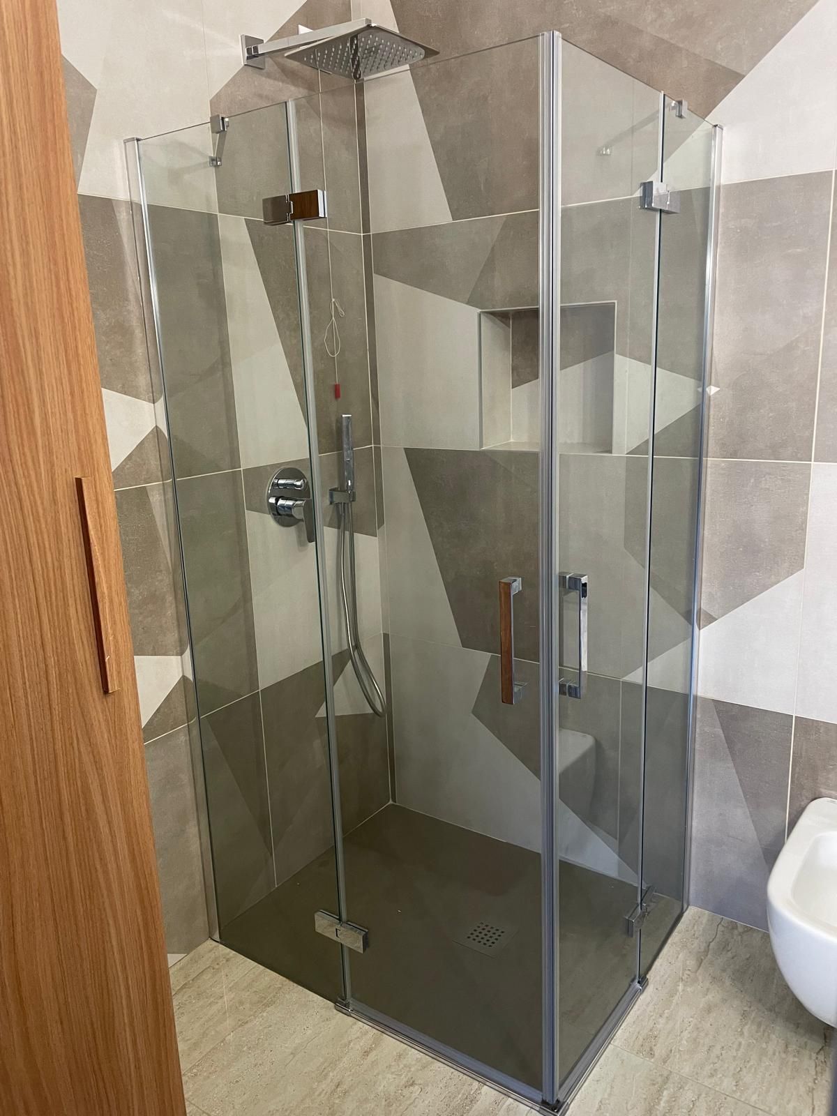 Box doccia angolare in vetro in un bagno piastrellato con porta in legno e WC visibile accanto.