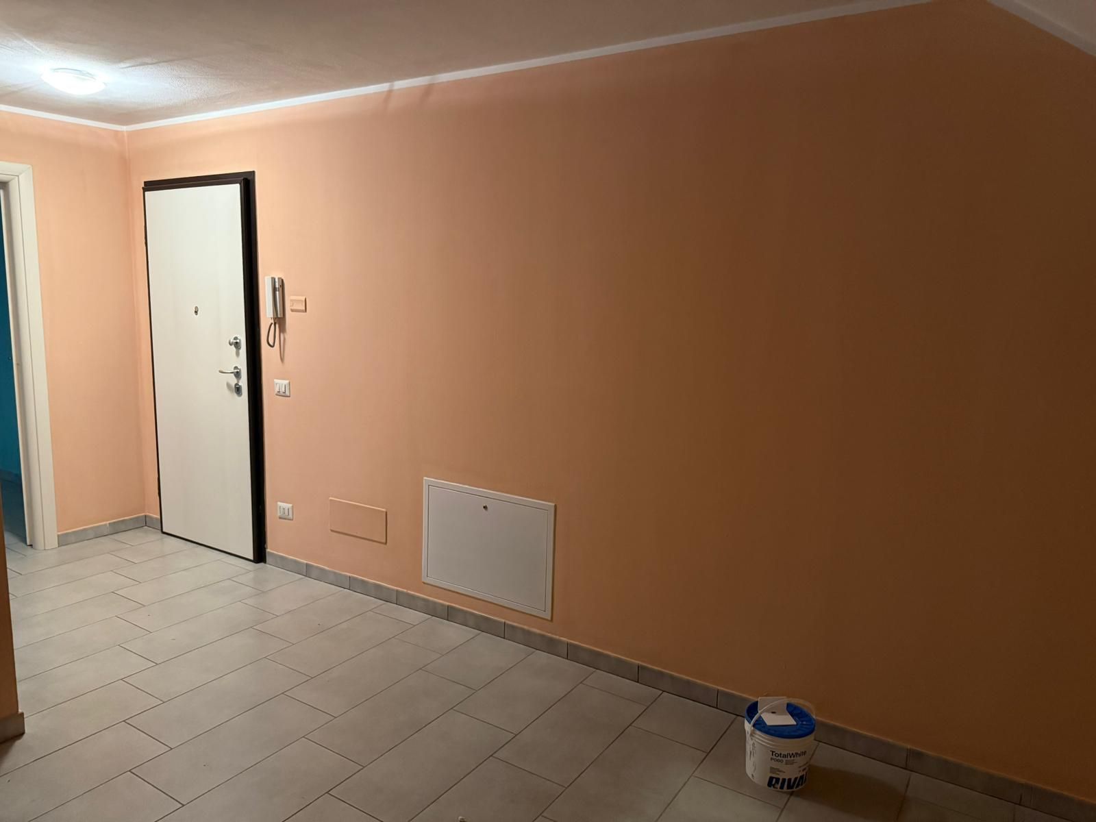 Corridoio vuoto color pesca con pavimento piastrellato, porte bianche e un secchio di vernice vicino al muro.