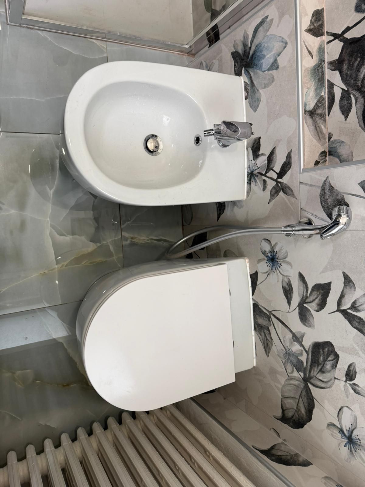 Piccolo bagno con lavabo bianco e WC sospeso accanto a piastrelle floreali e un termosifone.