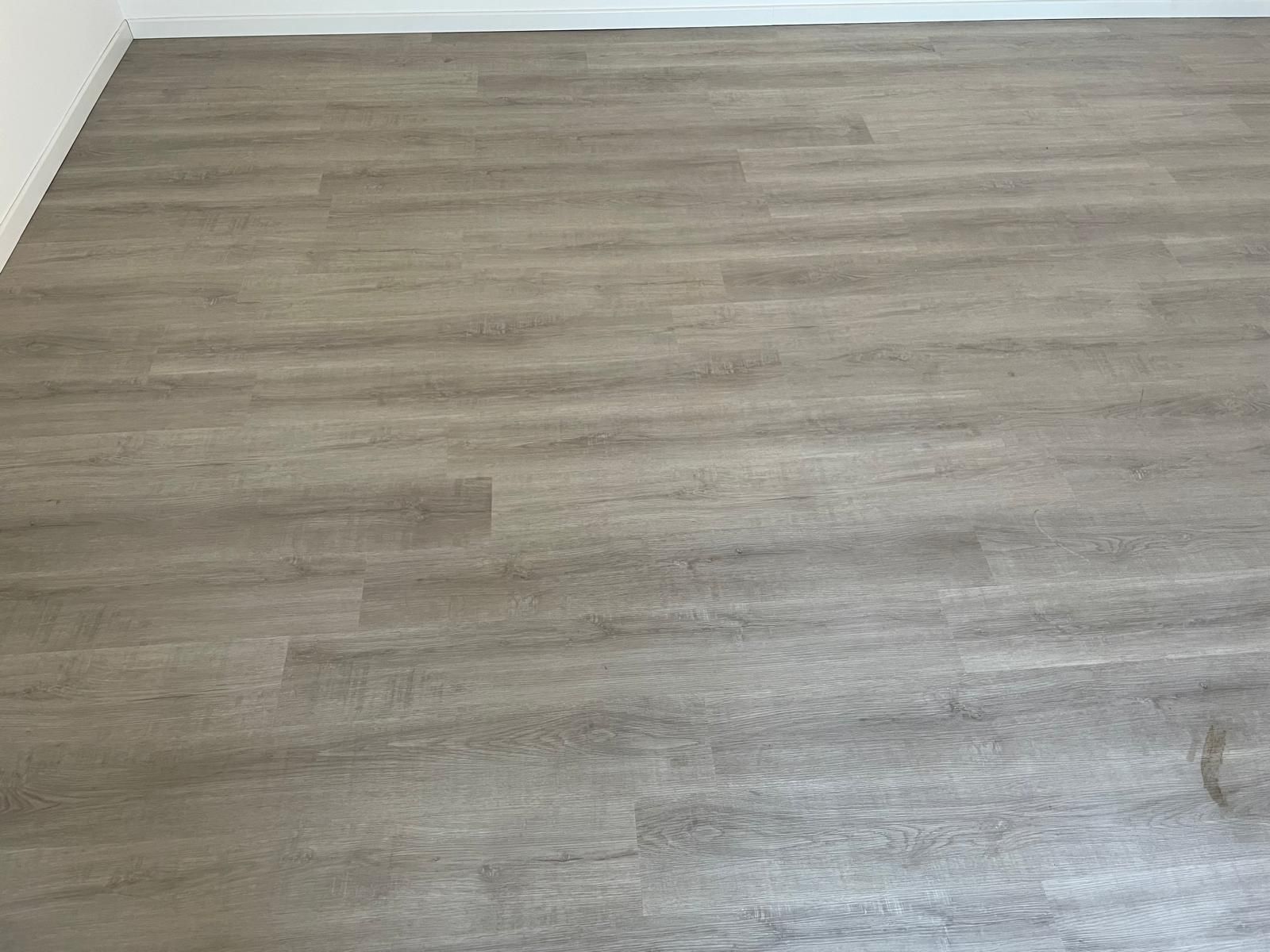 Stanza vuota con pavimento grigio effetto legno e battiscopa bianchi su una parete.