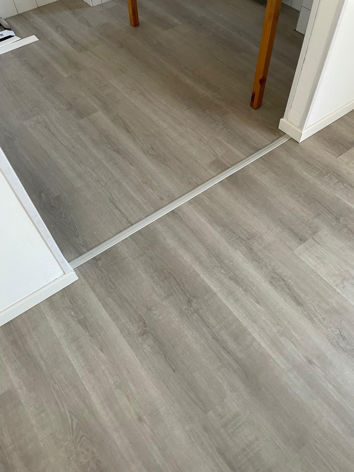 Pavimento in legno con una fascia bianca di transizione tra due stanze, e parte di un tavolo e una sedia visibili.