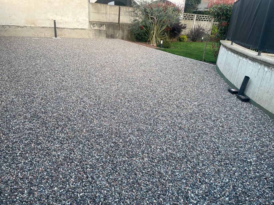 Patio posteriore ricoperto di ghiaia accanto a una casa, con un piccolo giardino e una recinzione sullo sfondo.