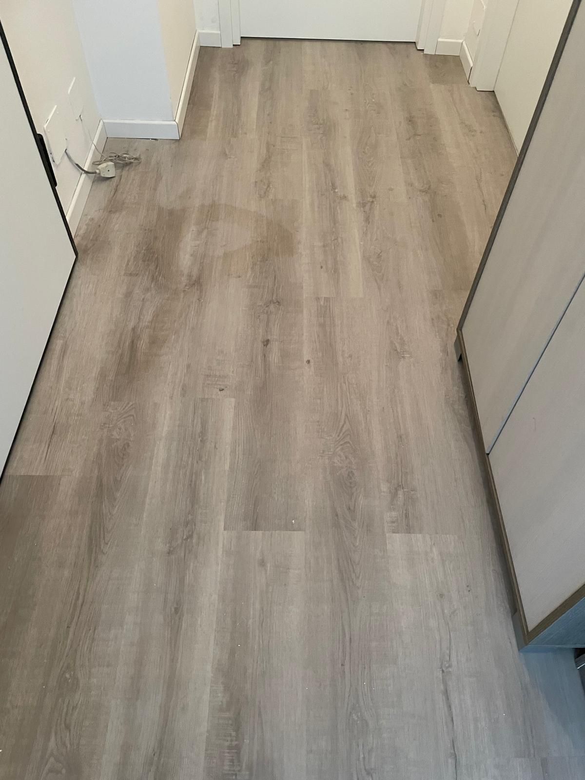 Corridoio vuoto con pavimento in legno chiaro e pareti bianche, visto verso una porta chiusa.