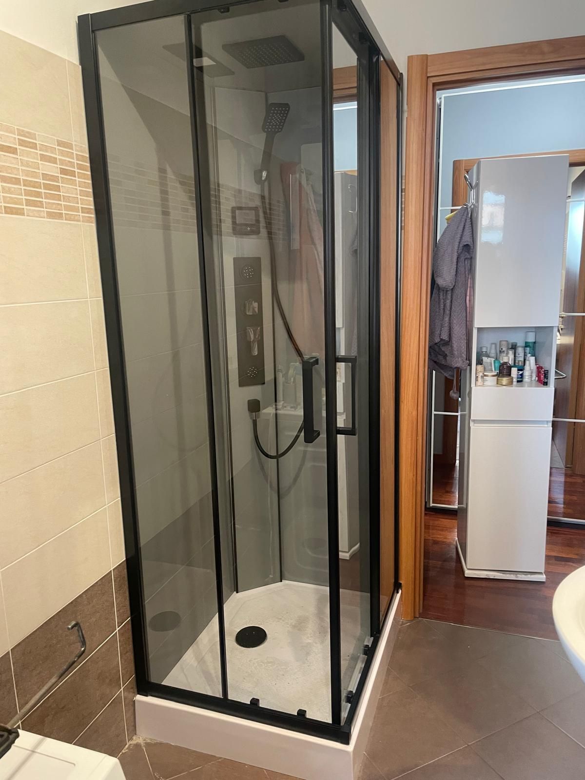 Doccia ad angolo con porta in vetro con cornice nera in un bagno, accanto a una porta aperta e a un mobiletto con specchio.