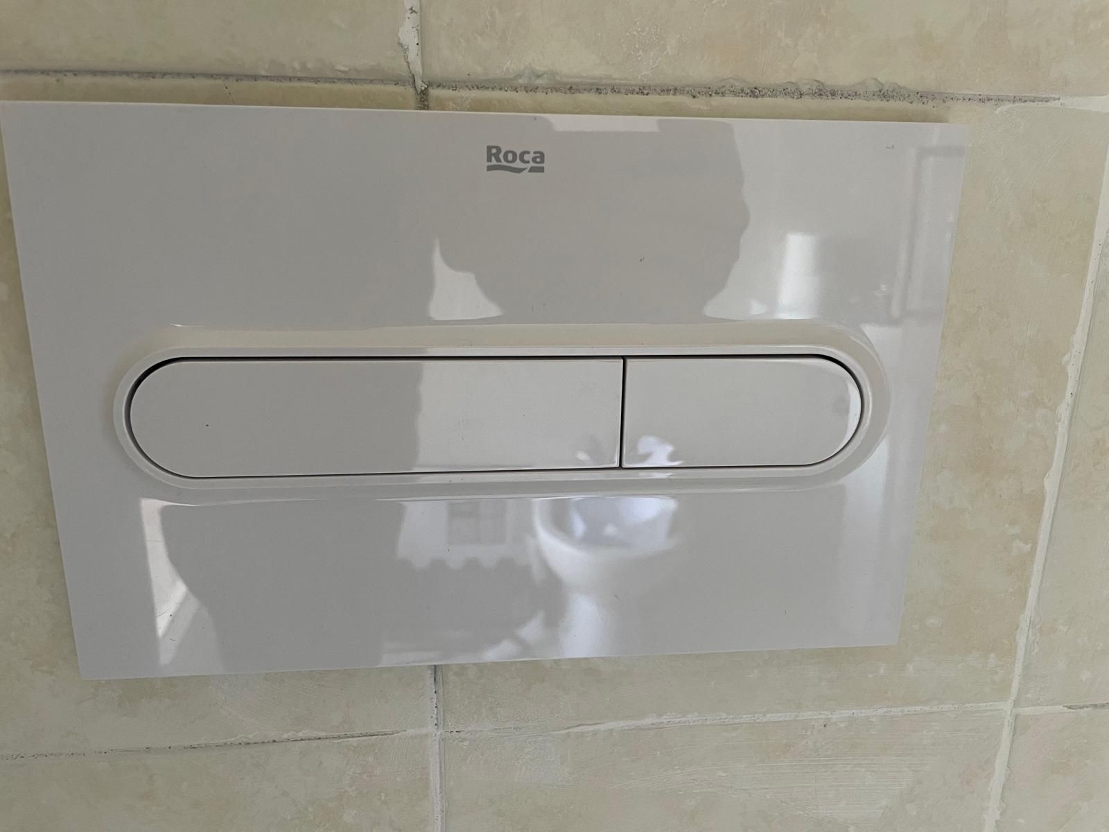 Pulsante di scarico doppio bianco Roca per WC su una parete piastrellata beige.