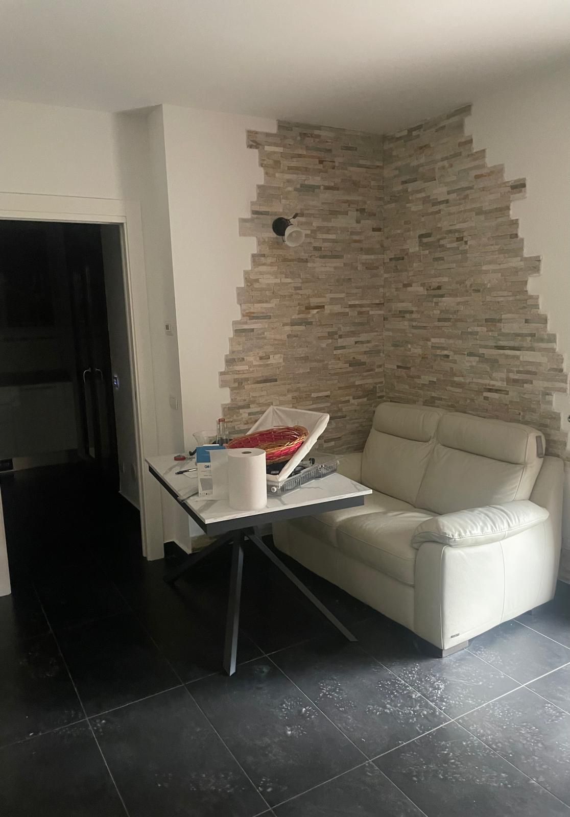 Piccolo soggiorno con divano bianco, tavolo ingombrato, parete con rivestimento in pietra e pavimento piastrellato scuro.